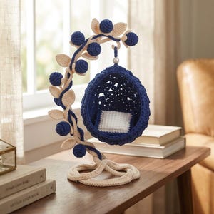 Puede incluir: Un adorno de silla colgante de ganchillo con un asiento azul oscuro en forma de huevo y un cojín cuadrado blanco, suspendido de un soporte curvo color crema decorado con bayas azul oscuro y hojas color crema, todo hecho de hilo.