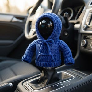 Op de afbeelding: Een blauwe gebreide hoodie-hoes voor een versnellingspook in een auto. Het kledingstuk heeft een capuchon, lange mouwen met geribbelde boorden, een kangoeroezak aan de voorzijde en een trekkoord met een strik. De stof heeft een dik breipatroon.