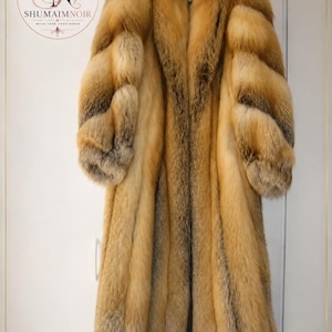 Peut inclure: Un long manteau en fourrure épaisse et texturée aux nuances de brun doré, de beige et de gris. Le vêtement présente des manches longues, un large col et un aspect dense et superposé. Un logo circulaire avec le texte SHUMAIMNOIR est visible.