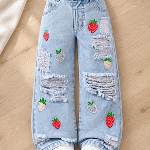 May include: Light blue denim jeans featuring a wide-leg silhouette, an elasticized waistband, and multiple distressed, frayed patches. The jeans are decorated with several small strawberry graphics in red and light pink colors, paired with white sneakers.