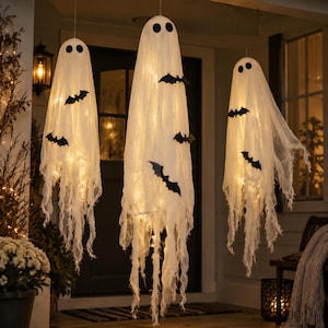 May include: Three hanging ghost decorations made of white, semi-sheer fabric with frayed edges. Each ghost features two black circular eyes and is adorned with several black bat-shaped cutouts. The figures are illuminated from within by lights.