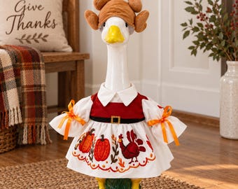 May include: A white goose figurine wearing a brown plush turkey hat and a white dress with a red bodice, black belt, and orange ribbon sleeve ties. The skirt features embroidered orange pumpkins and a red turkey with green leaf accents.