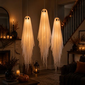 Puede incluir: Tres decoraciones de fantasmas colgantes hechas de tela blanca transparente con ojos circulares negros. Cada fantasma está iluminado desde el interior por una luz cálida, creando un efecto brillante. La tela cae con bordes deshilachados.