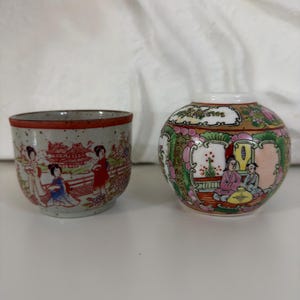 Puede incluir: Una taza de té de cerámica con acabado gris moteado y borde rojo, que presenta una escena pintada de figuras en un paisaje, junto a un pequeño tarro de porcelana redondo y multicolor decorado con intrincados motivos florales y figuras humanas.