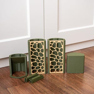 Könnte beinhalten: Ein Set aus olivgrünen Moss Buddy-Komponenten, bestehend aus zwei hohen vertikalen Strukturen mit beigen giraffenartigen Mustern, einem kurzen Teil mit offener Basis, einer massiven Kappe mit der Aufschrift Moss Buddy und mehreren kleinen Verbindungsstäben.