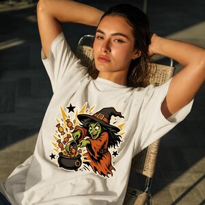 May include: A white t-shirt featuring a graphic illustration of a green-skinned witch wearing a black hat and orange robe. The witch is stirring a black cauldron with orange candies flying out, surrounded by yellow starbursts and black stars.