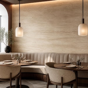 May include: A dining area featuring two round wooden tables with beige upholstered chairs and a curved beige banquette. Two white ribbed pendant lights hang from the ceiling above the tables, which are set with wine glasses and white plates.