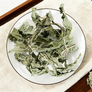 May include: A collection of dried, crinkled green leaves with visible stems arranged on a round white plate. The leaves exhibit a muted, sage-like colour with a brittle, dehydrated texture, showcasing natural veins and curled edges throughout the pile.