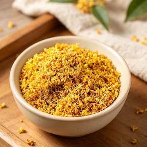 May include: A round, off-white ceramic bowl filled with a generous portion of dried, vibrant yellow osmanthus flowers. The small, delicate blossoms are densely packed, showcasing their natural golden-orange hues and intricate, curled petal textures.