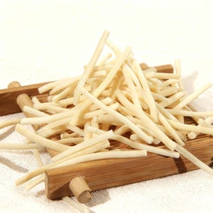 Pode incluir: Um monte de palitos alimentares cilíndricos, finos e de cor esbranquiçada, dispostos sobre uma bandeja retangular de ripas de bambu. Os palitos são uniformes em forma e cor, empilhados de forma solta sobre a superfície de madeira.