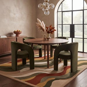 May include: A round wooden dining table with four olive green velvet chairs featuring curved armrests, placed on a rectangular area rug with a wavy pattern in shades of rust, olive, mustard, and beige, set in a room with a large arched window.