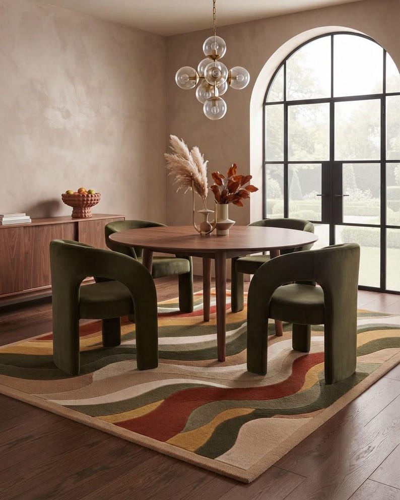 May include: A round wooden dining table with four olive green velvet chairs featuring curved armrests, placed on a rectangular area rug with a wavy pattern in shades of rust, olive, mustard, and beige, set in a room with a large arched window.