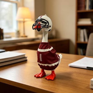 May include: A white goose figurine wearing a maroon football jersey with a white lace graphic and a white football helmet with a black face mask. The goose has bright orange feet and an orange beak, standing upright as a decorative tabletop ornament.
