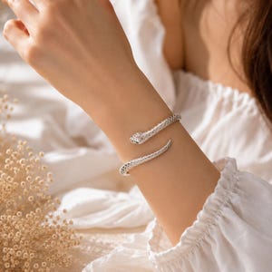 以下が含まれることがあります： とぐろを巻いたヘビの形をしたシルバーカラーの金属製ブレスレット。ヘビの全長にわたって鱗のような質感のパターンが施されており、一方の端には明確な頭部、もう一方の端には先細りの尾があり、手首に巻き付くデザインになっています。