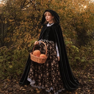 May include: A black hooded velvet cloak worn over a white collared shirt and a long dark skirt with a floral pattern and white lace trim at the hem. A woven wicker basket containing two orange pumpkins is held in front of the skirt.