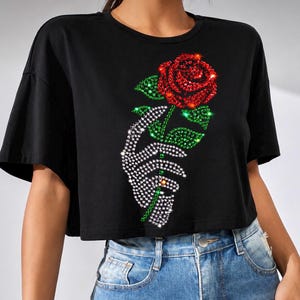 May include: A black short-sleeved crop top featuring a graphic design made of sparkling rhinestones. The design depicts a hand holding a single red rose with green leaves. The rhinestones are red, green, and clear, creating a shimmering effect.