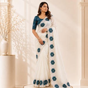 May include: A white saree featuring a dark teal blouse with a geometric pattern and a matching teal floral border applique along the edge of the fabric. The AaviraCouture logo and text are visible at the top center of the image above the garment.