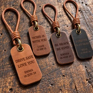 May include: Four rectangular leather keychains with rounded corners, each featuring a leather loop attached by a brass ring. The keychains are engraved with custom text including names and dates, such as DRIVE SAFE LOVE YOU, HOME IS WITH YOU, BE BRAVE BE KIND, and FOREVER IN MY HEART.