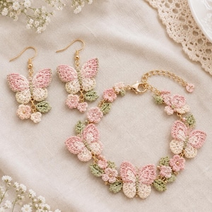 Può includere: Un set di gioielli composto da due orecchini pendenti e un bracciale con farfalle all'uncinetto nei toni del rosa e del crema, arricchiti da piccole foglie verdi e fiori rosa, collegati da componenti e catenelle in metallo dorato.