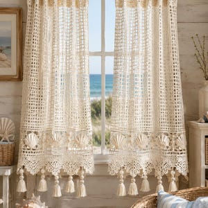 May include: A pair of off-white crochet curtains hanging from a wooden rod. The curtains feature a grid-like pattern, a decorative border with integrated seashell motifs, and a bottom edge finished with dangling tassels and small round beads.