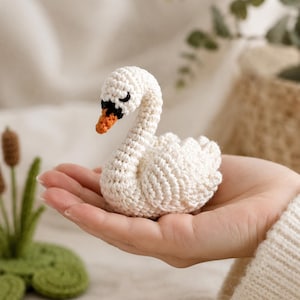 Puede incluir: Una pequeña figura de cisne tejida a mano en ganchillo con hilo color blanco roto. El cisne presenta un cuello curvado, detalles de alas texturizadas y un pico de color negro y naranja. La pieza está construida con puntos visibles en todo el cuerpo.