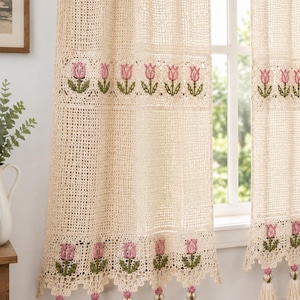 May include: A pair of cream-coloured crochet curtains featuring a grid-like pattern and horizontal bands of embroidered pink tulips with green stems and leaves. The bottom edges are finished with a scalloped border and hanging cream-coloured tassels.