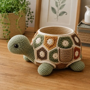 Könnte beinhalten: Ein gehäkelter Schildkrötenkorb mit einem Panzer aus mehrfarbigen sechseckigen Motiven in Salbeigrün, Senfgelb, Creme und Braun. Die Schildkröte hat einen hellgrünen Kopf mit einem schwarzen Auge und vier dazu passende grüne Beine.