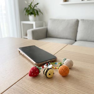 May include: A smartphone charm featuring four small crocheted figures: a red strawberry with white dots, a yellow and black striped bee with white wings, an orange sphere, and a beige sphere, all connected by colorful beads to a metal loop.