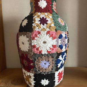 May include: A decorative vase covered in a multi-colored crochet granny square cozy. The textile cover features various square patterns in shades of red, white, black, green, blue, yellow, and tan, tightly fitted around the rounded ceramic vessel.