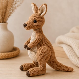 Puede incluir: Un juguete de canguro tejido a ganchillo con cuerpo marrón claro, vientre y parte interior de las orejas en color crema. El peluche tiene detalles en marrón oscuro en la nariz, manos y pies, además de ojos negros de plástico y una cola.