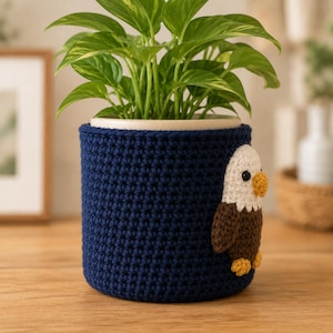 Op de afbeelding: Een donkerblauwe gehaakte plantenpotcover met een driedimensionale applicatie van een Amerikaanse zeearend aan de zijkant. De adelaar heeft een witte kop, bruin lichaam, gele snavel en gele poten. Een groene plant staat in de cilindrische hoes.