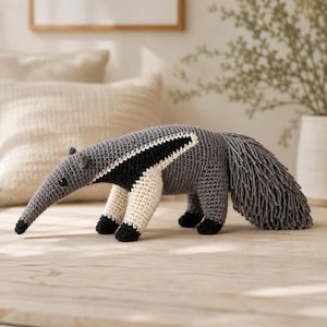 Puede incluir: Un juguete de oso hormiguero tejido a ganchillo hecho a mano con un hocico largo, cuerpo gris con un patrón de rayas diagonales en blanco y negro, y una cola gris texturizada y peluda. La figura se sostiene sobre cuatro patas con pies negros.