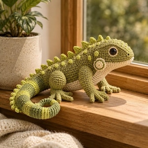 May include: A crocheted iguana toy featuring a textured green body with lighter green stripes on the tail, a cream-colored underbelly, a row of small spikes along the back, and large dark brown eyes. The stuffed animal is crafted with visible yarn stitches.