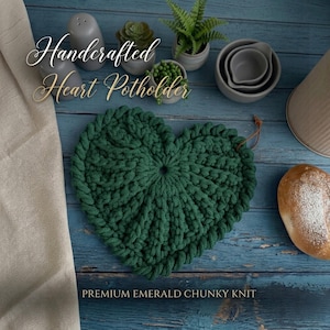 May include: A dark green heart-shaped chunky knit potholder with a small leather loop. The text "Handcrafted Heart Potholder" and "PREMIUM EMERALD CHUNKY KNIT" is visible. The item is surrounded by small potted plants, grey bowls, and a loaf of bread.