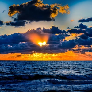 May include: A vibrant sunset over the ocean featuring a bright orange and yellow light breaking through dark, heavy clouds. The horizon line separates the deep blue sea with gentle waves from the dramatic sky filled with golden light rays.