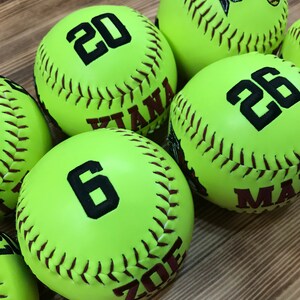 May include: Six yellow softballs with black numbers and brown stitching. The numbers on the softballs are 20, 26, 6, and the names "Kiana", "Zoe", and "Max" are written on the softballs.