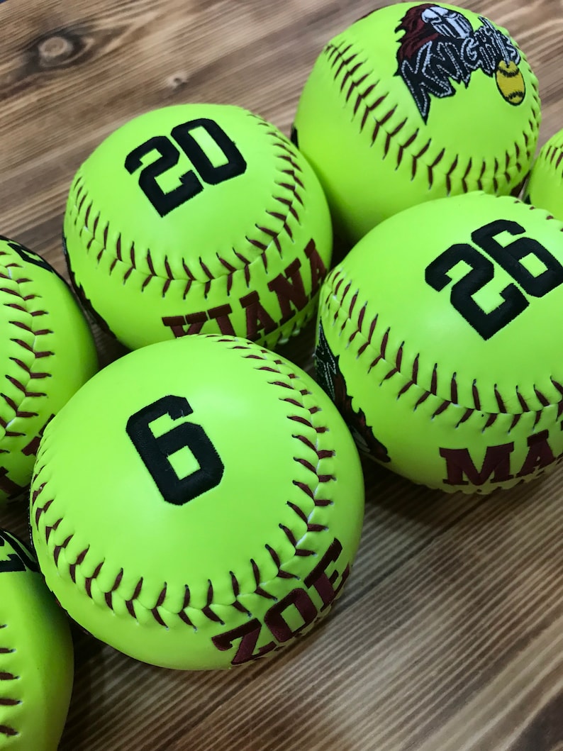 May include: Six yellow softballs with black numbers and brown stitching. The numbers on the softballs are 20, 26, 6, and the names "Kiana", "Zoe", and "Max" are written on the softballs.