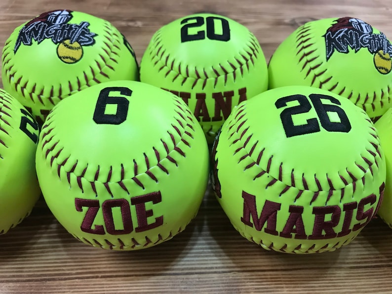 May include: Six lime green softball with maroon stitching. The softballs are personalized with names and numbers. The names are Zoe, Maris, and the numbers are 6, 20, and 26.