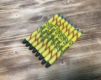 May include: Eight yellow and red softball-themed pens with black glitter. Each pen has a name and number printed on it. The names are Colbie, Aria, Brooklynn, Belle, Hia, Leah, Ella, and Avery.