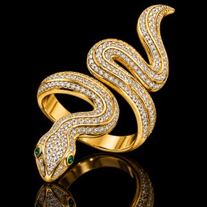 May include: A gold ring shaped like a coiled snake, featuring a pavé-set surface of numerous small clear stones covering the body. The snake head is accented with two small, round, vibrant green stones for eyes, creating a detailed, reflective piece.