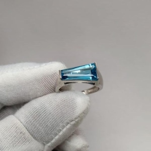 Könnte beinhalten: Ein silberfarbener Ring mit einem einzelnen, länglichen, trapezförmig geschliffenen blauen Edelstein, der horizontal eingesetzt ist. Die Ringschiene ist glatt und metallisch und bildet einen klaren Rahmen für den facettierten, blauen Stein.