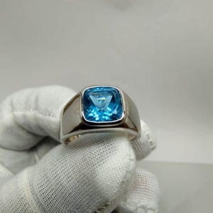 Könnte beinhalten: Ein silberfarbener Ring mit einem großen, quadratisch geschliffenen, leuchtend blauen Edelstein, der in ein glattes, poliertes Metallband gefasst ist. Der Stein hat eine facettierte Oberfläche, die das Licht reflektiert, und das Band ist breit.