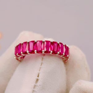 Könnte beinhalten: Ein silberfarbener Eternity-Ring mit einer durchgehenden Reihe von leuchtenden, rechteckig geschliffenen, tief rosa-roten Edelsteinen in einer Krappenfassung. Der Ring ist vor einem neutralen, weichen Hintergrund abgebildet.