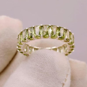 Könnte beinhalten: Ein silberner Eternity-Ring mit einer durchgehenden Reihe von rechteckigen, facettierten, hellgrünen Peridot-Edelsteinen, die in einen polierten Metallrahmen gefasst sind. Der Ring wird vor einem neutralen Hintergrund gezeigt.