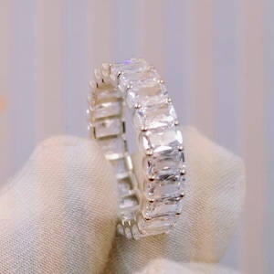 Könnte beinhalten: Ein Eternity-Ring mit einer durchgehenden Reihe klarer, rechteckiger Steine im Smaragdschliff, die in ein poliertes, silberfarbenes Metallband gefasst sind. Die Steine sind nebeneinander angeordnet und erzeugen ein gleichmäßiges, funkelndes Erscheinungsbild.