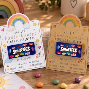 Könnte beinhalten: Zwei rechteckige Geschenkkarten mit Regenbogen-Grafik und dem Text AUF EIN kunterbuntes KINDERGARTENJAHR MIT GANZ VIEL. Jede Karte enthält eine kleine blaue Schachtel Smarties mini. Die Karten sind mit bunten Symbolen wie Sternen, Herzen und einer Rakete verziert.