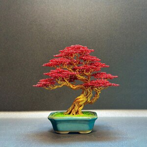 Puede incluir: Una escultura de árbol bonsái hecha de alambre con un tronco y ramas retorcidas de color dorado y follaje de alambre rojo vibrante. La escultura está colocada en una pequeña maceta de cerámica rectangular de color verde azulado con musgo.