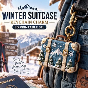 May include: A blue suitcase-shaped keychain charm featuring a relief design of a snow-capped mountain, evergreen trees, and snowflakes. The charm has cream-colored straps with gold buckles and is attached to a gold-toned metal lobster clasp.
