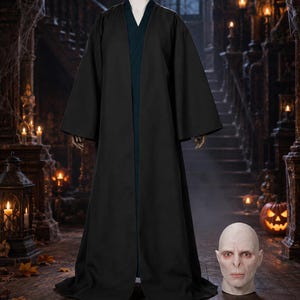 May include: A floor-length black robe with wide sleeves and a dark teal inner layer, displayed on a mannequin torso. Beside the robe sits a pale, hairless, human-like mask with a flat nose and prominent brow, resembling a fictional character.