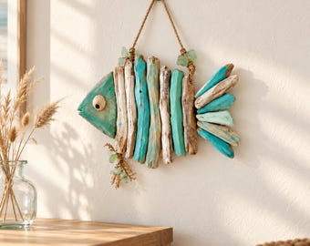 May include: A wall hanging shaped like a fish, constructed from vertical pieces of driftwood in natural tan and painted turquoise hues. The fish features a single round eye, a rope hanger at the top, and small green sea glass accents.